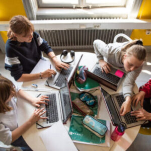 Four children gather around laptops in a classroom helping each other