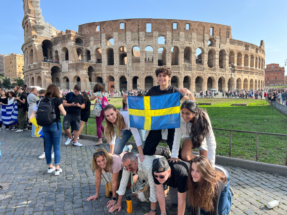 Students pose in front of the coliseum with a Swedish flag